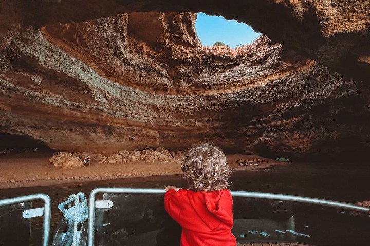 Child in red hoodie views cave from boat with light shining through opening above.