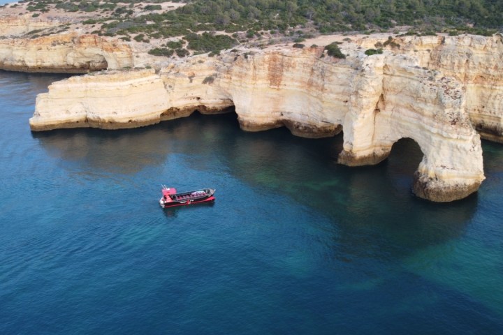 Red boat near coastal cliffs with arch formation in calm blue water.