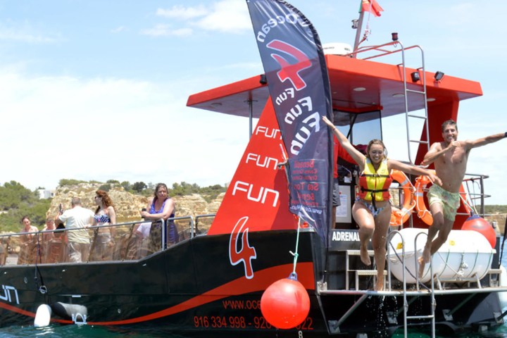 Two people jump off a boat with fun banners, others watch onboard near rocky coastline.