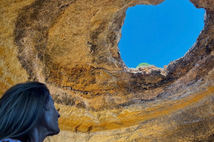Person gazing up at a rock cave with large circular opening to the sky.