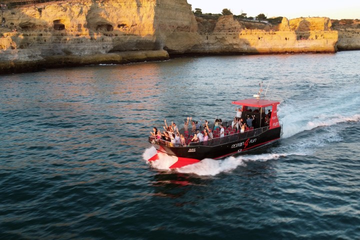 Red and black boat with people cruising near rocky cliffs at sunset.