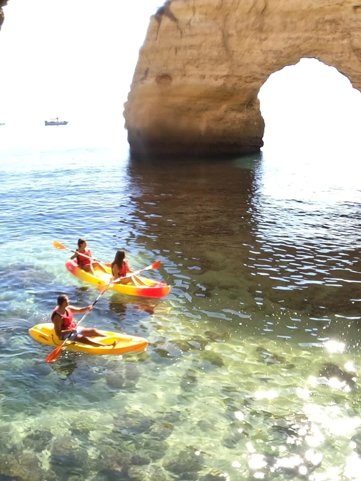 Three kayakers in orange kayaks near a large coastal rock arch.
