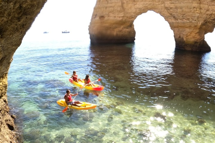 Three kayakers in orange kayaks near a large coastal rock arch.