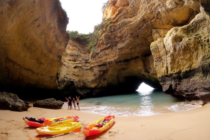 People with kayaks on a sandy beach inside a large cave.