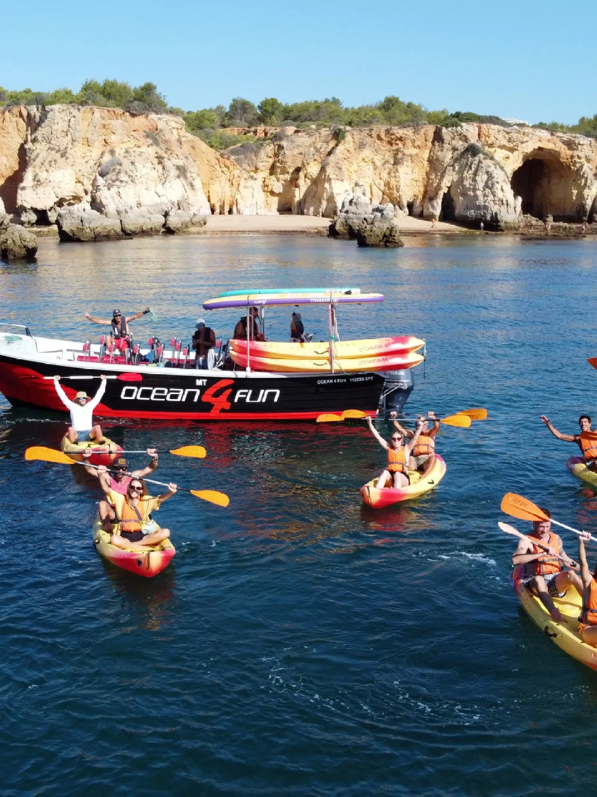 Group kayaking near a tour boat by rocky shore and cliffs.