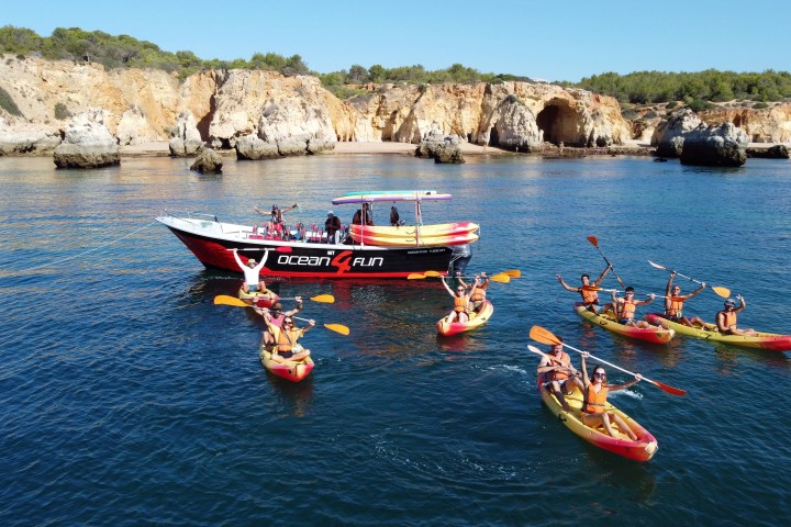 Group kayaking near a tour boat by rocky shore and cliffs.