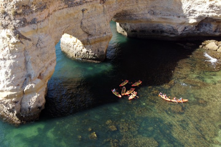 Kayakers paddle near large coastal rock arch over clear water.