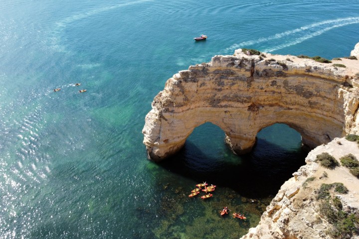 Rock formation with natural arches over clear blue ocean, small boats nearby.