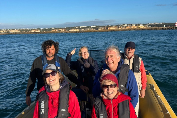 Group of six people in life jackets on a small boat with a coastal backdrop.