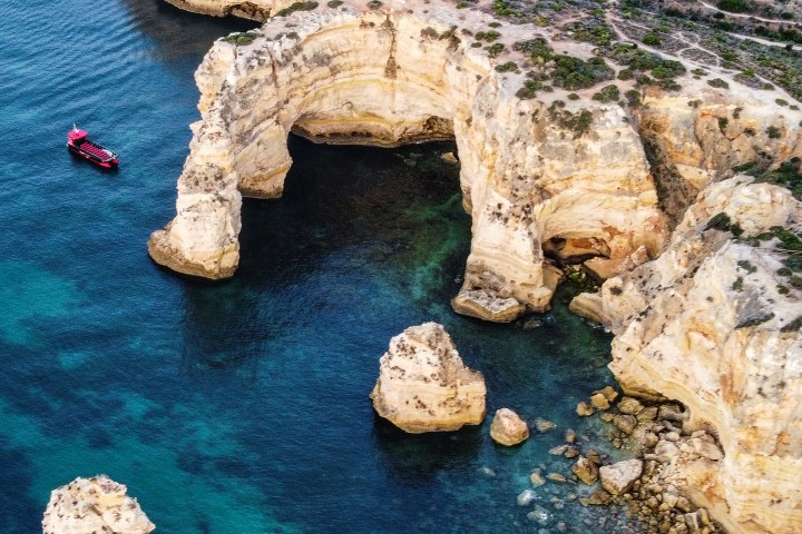 Aerial view of rocky coastal cliffs with a red boat in blue-green water.