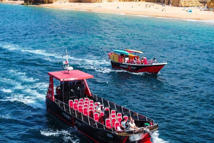 Two tourist boats on blue water near a rocky coastline with a sandy beach.