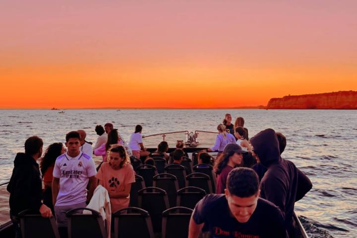 People on a boat at sunset with orange sky and distant cliffs.
