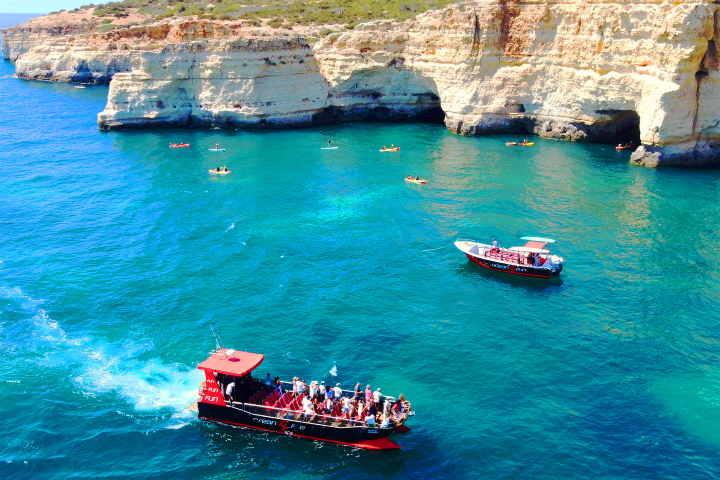 Boats with tourists in clear blue water near rocky cliffs and kayakers.