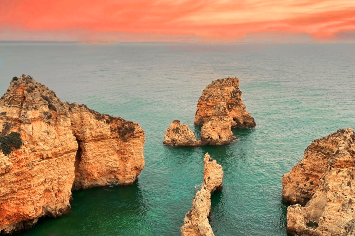 Rock formations in green sea under vibrant red-orange sunset sky.