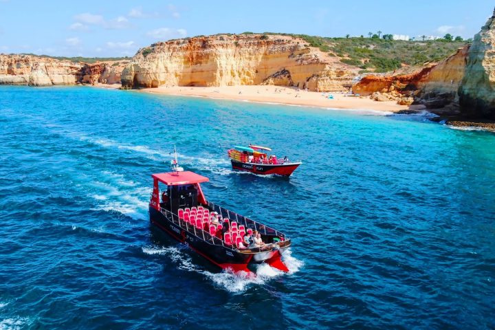 Two tour boats in a blue ocean near rocky cliffs and a sandy beach.