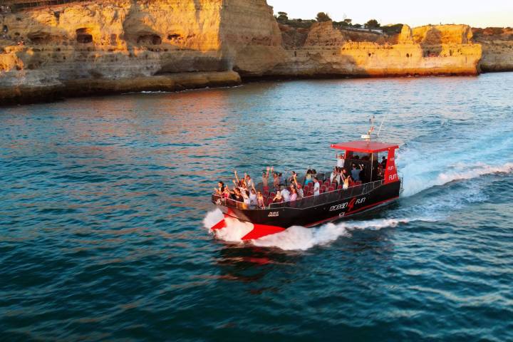 Tour boat with passengers on blue water near cliffs at sunset.