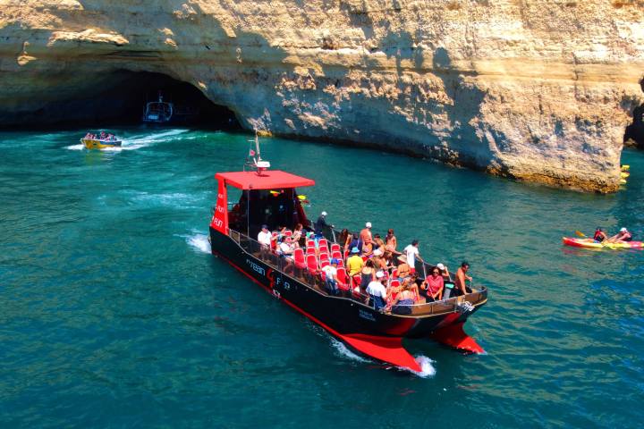 Tour boat with people near a sea cave entrance, clear blue water, and kayakers nearby.